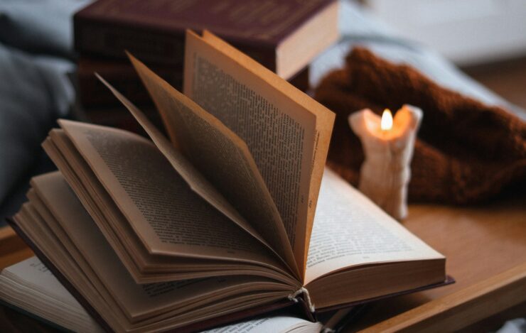 Photograph of two open books beside a candle. An additional stack of books and blankets or other cloth are visible in the background.