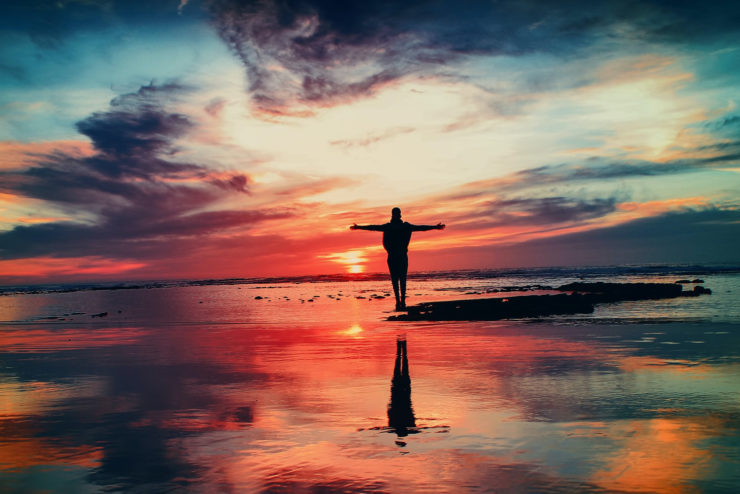 Photo of a sunset over a body of water, with colorful clouds. The silhouette of a human figure stands near the center of the frame with their arms outstretched.